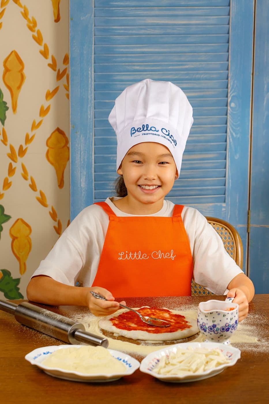 Девочка в поварском колпаке и оранжевом фартуке Little Chef делает пиццу