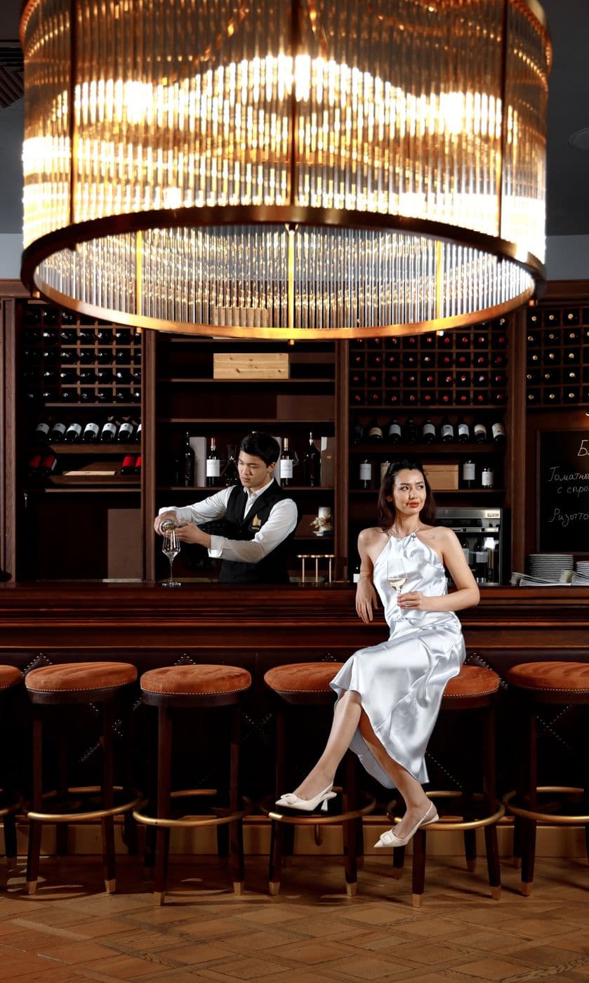 A bartender at the counter and a girl in a silver dress on a bar stool under a large chandelier