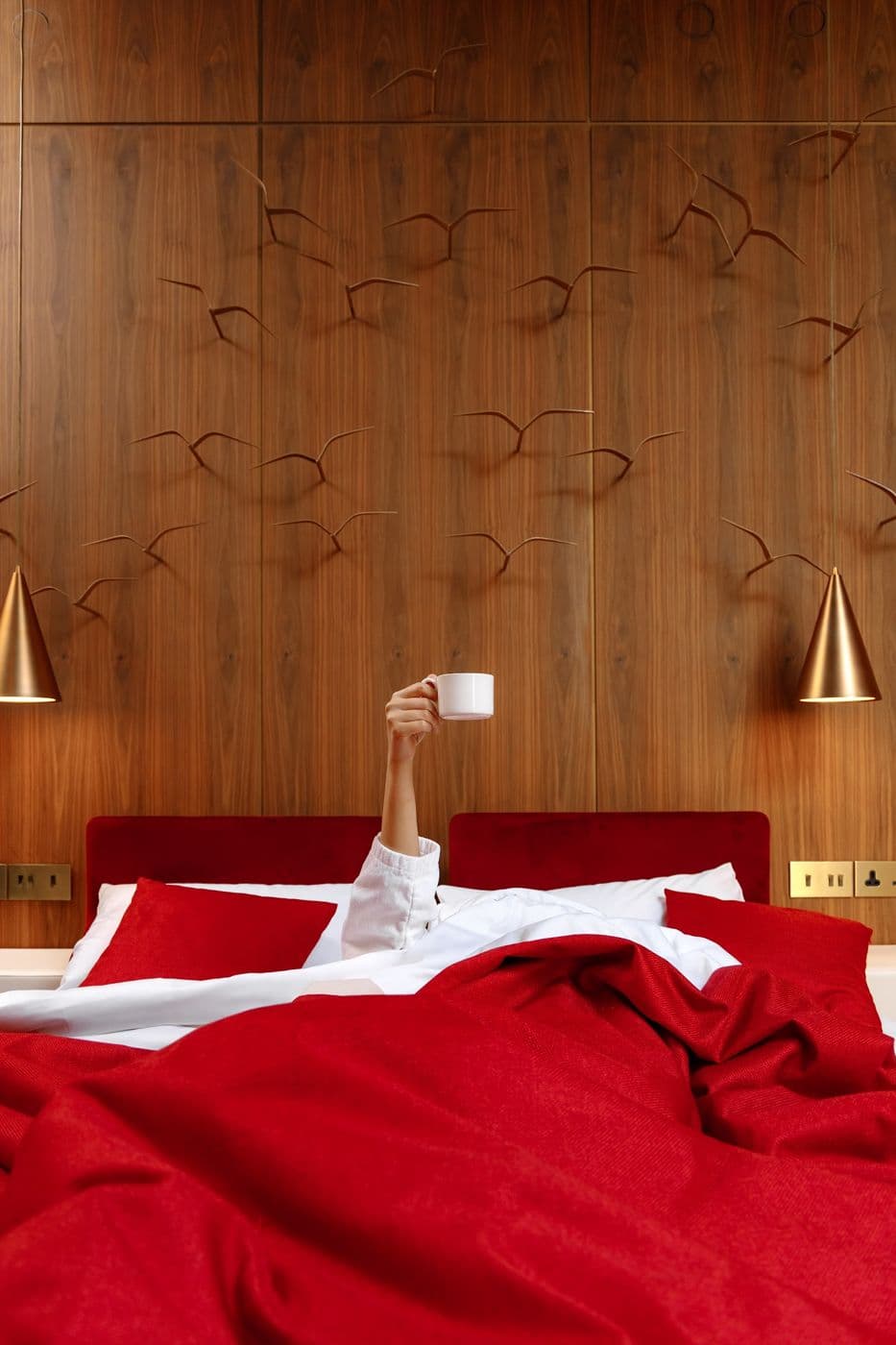 A hand with a cup coming out from under a red blanket on a bed in a room with wooden panels