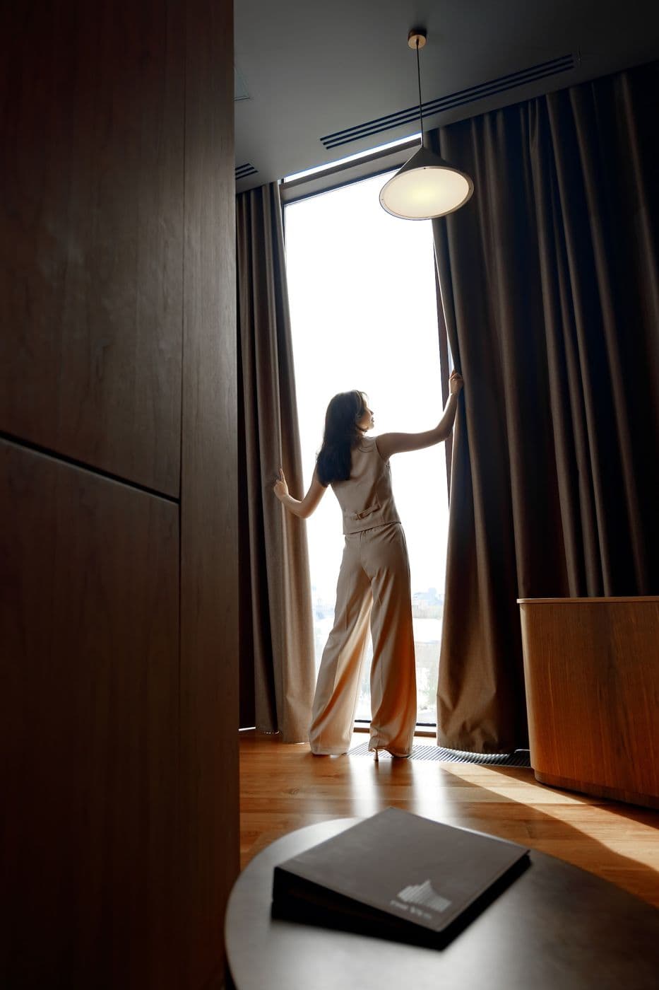 A girl by the window in a room with dark curtains, a closed folder on the table